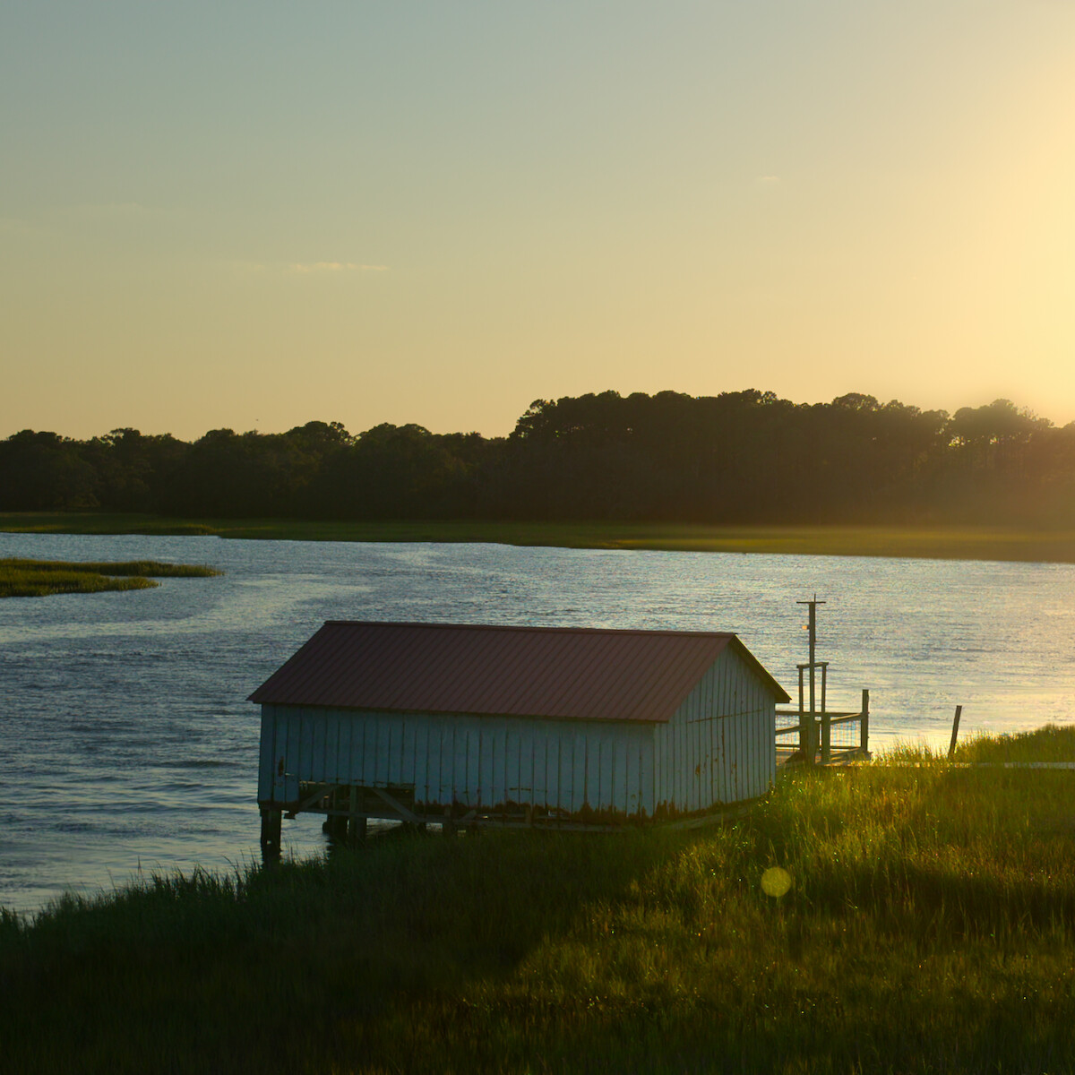 A wooden boathouse with a red roof sits on stilts over a calm river at sunset, surrounded by grassy banks and trees in the distance, bathed in warm golden light.