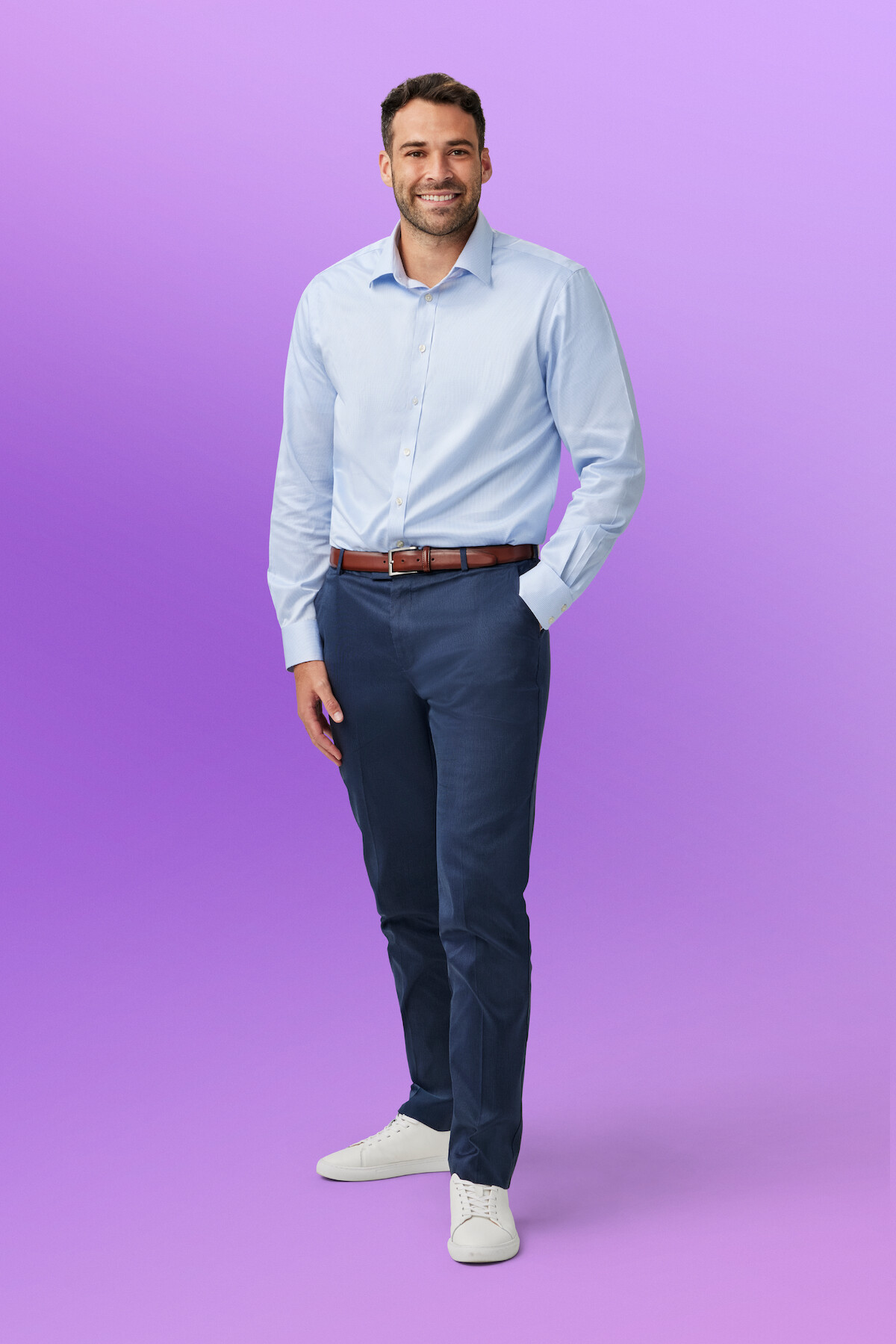 Alex H. in a light blue shirt, navy pants, and white sneakers standing and smiling against a purple gradient studio background.