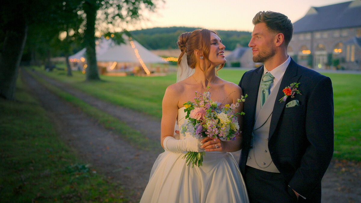 Bride and groom smiling at each other outdoors at sunset, bride holding bouquet, groom in suit, elegant wedding setting with tents and greenery, joyful and romantic atmosphere.