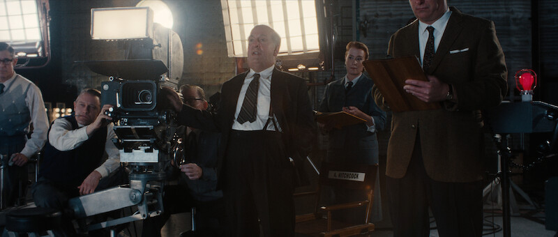 A man in a suit stands beside a large film camera on a movie set, surrounded by crew members and bright studio lights.