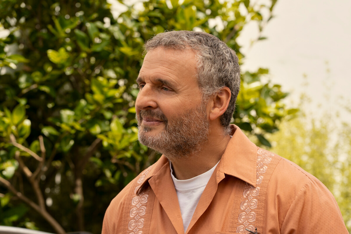 Middle-aged man with gray hair and beard wearing an orange shirt stands outdoors, smiling and looking to the side, with leafy green trees and a bright, natural background behind him.
