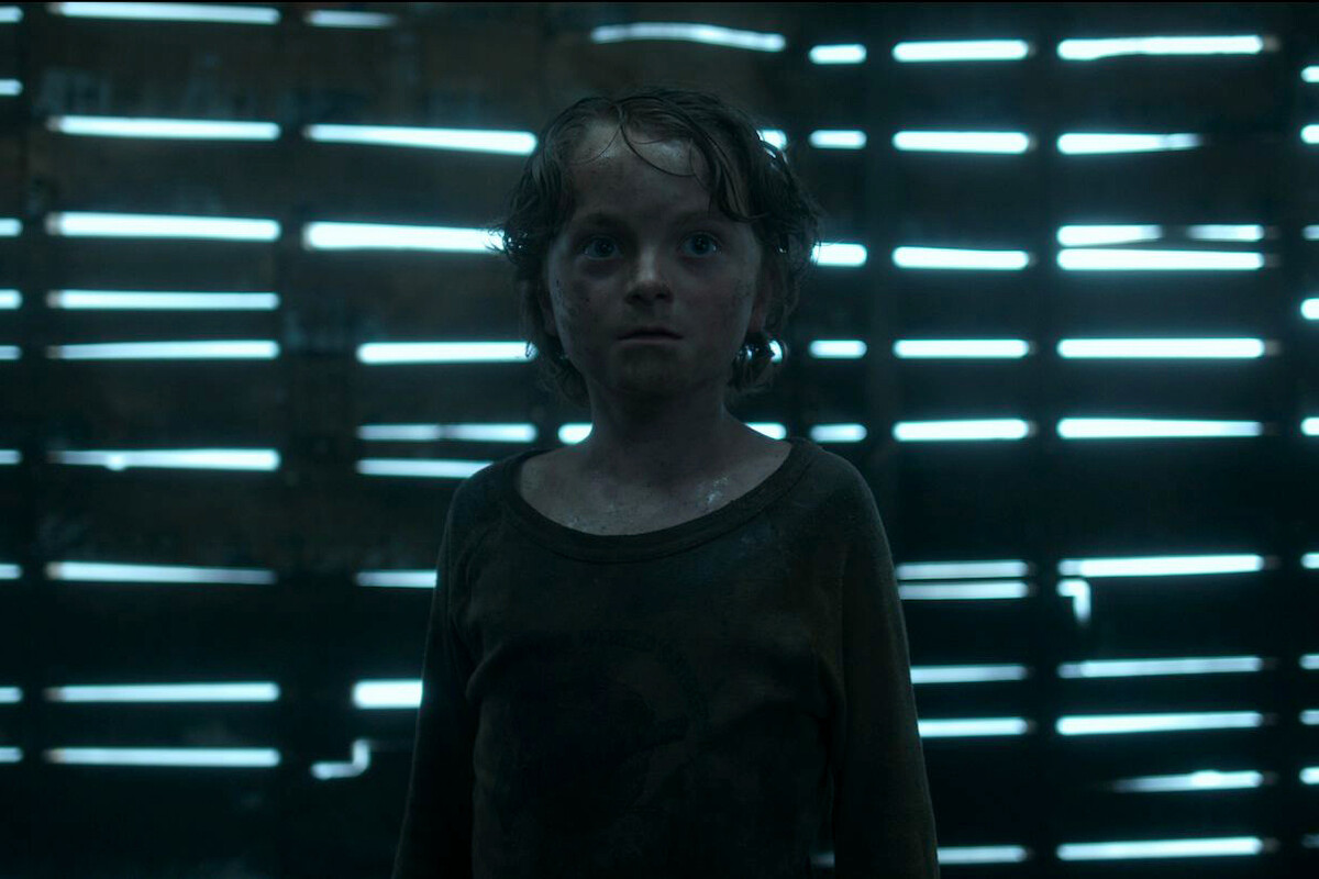 A young, wet child stands in a dimly lit, rustic wooden structure with slatted walls and beams of light shining through the gaps, creating an eerie and suspenseful atmosphere.