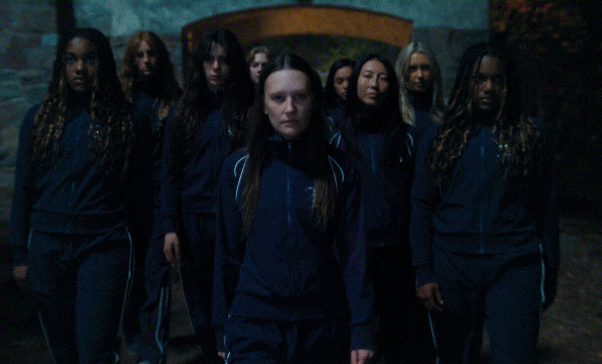 A group of serious-faced young women in matching navy tracksuits stand outdoors in low light, possibly at night, in front of a stone archway surrounded by trees.