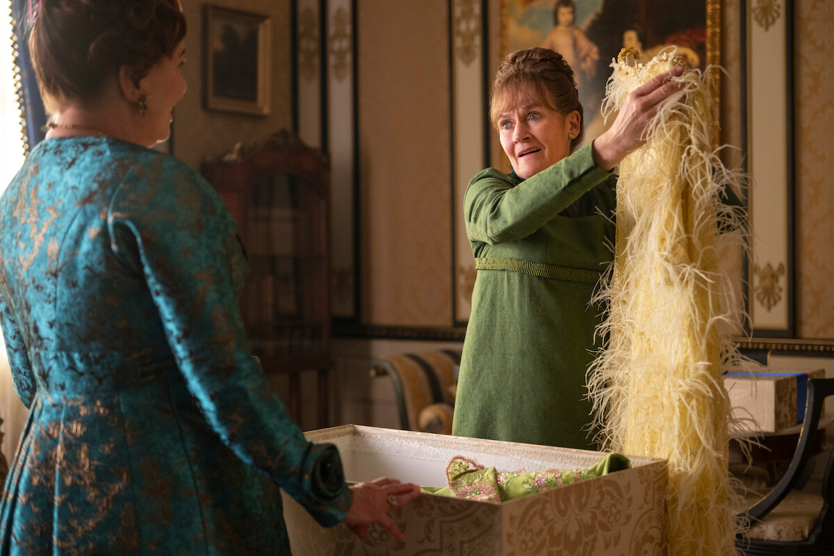 Two women in elegant period clothing are in a lavish, ornately decorated room; one woman is holding up a yellow feathered garment from a box, both appear engaged in conversation.