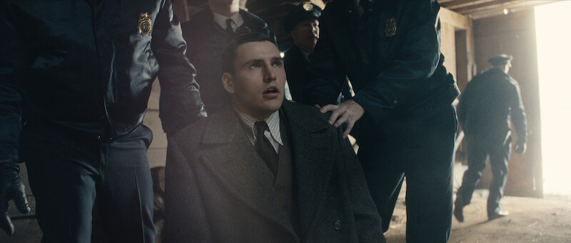 A young man in a suit coat is held by police officers, looking distressed as he is led out of a dimly lit building toward a bright exit.