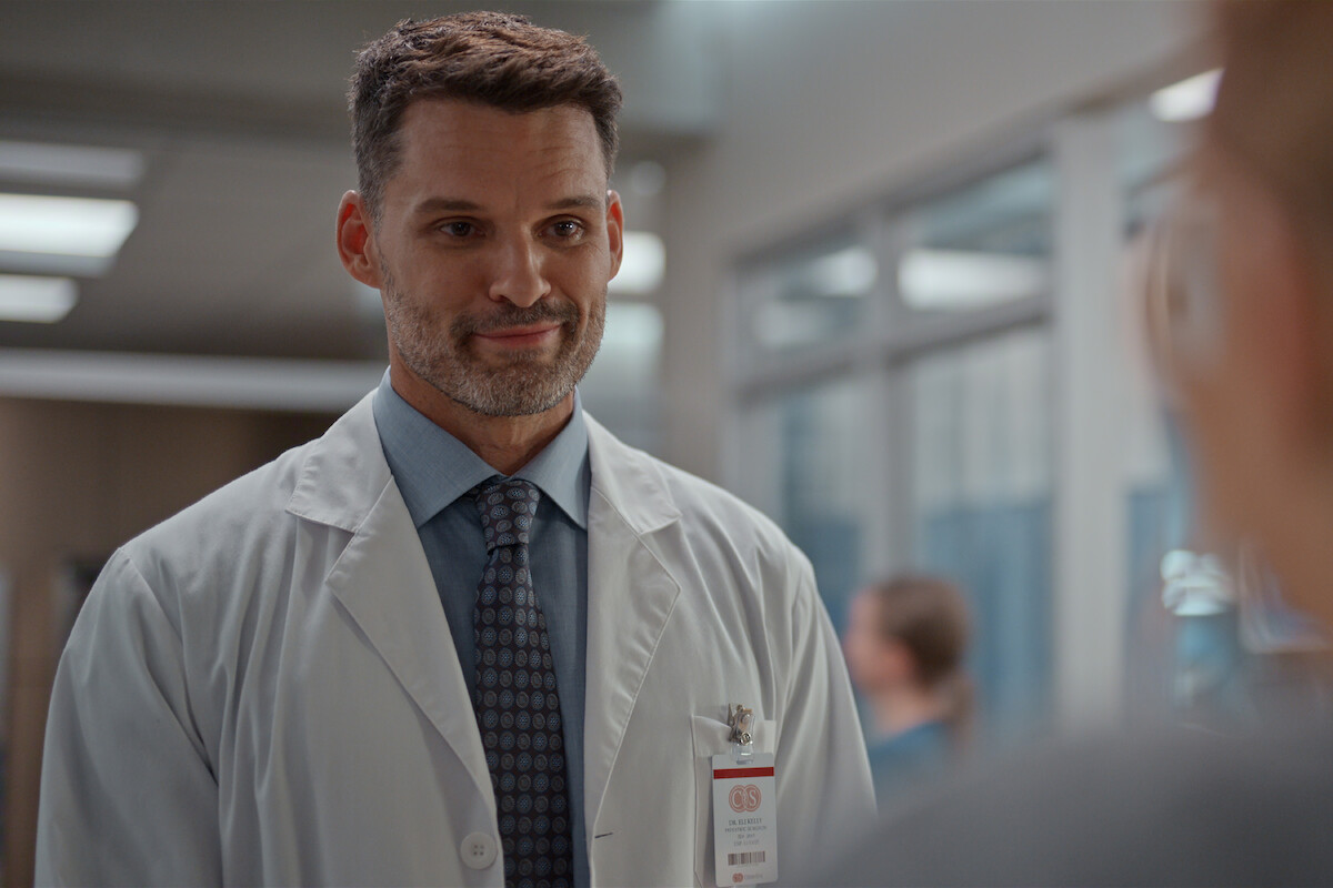 Male doctor in a white lab coat with a hospital ID badge, standing indoors in a modern medical facility, talking to a person out of focus in the foreground, with medical staff and equipment in the background.