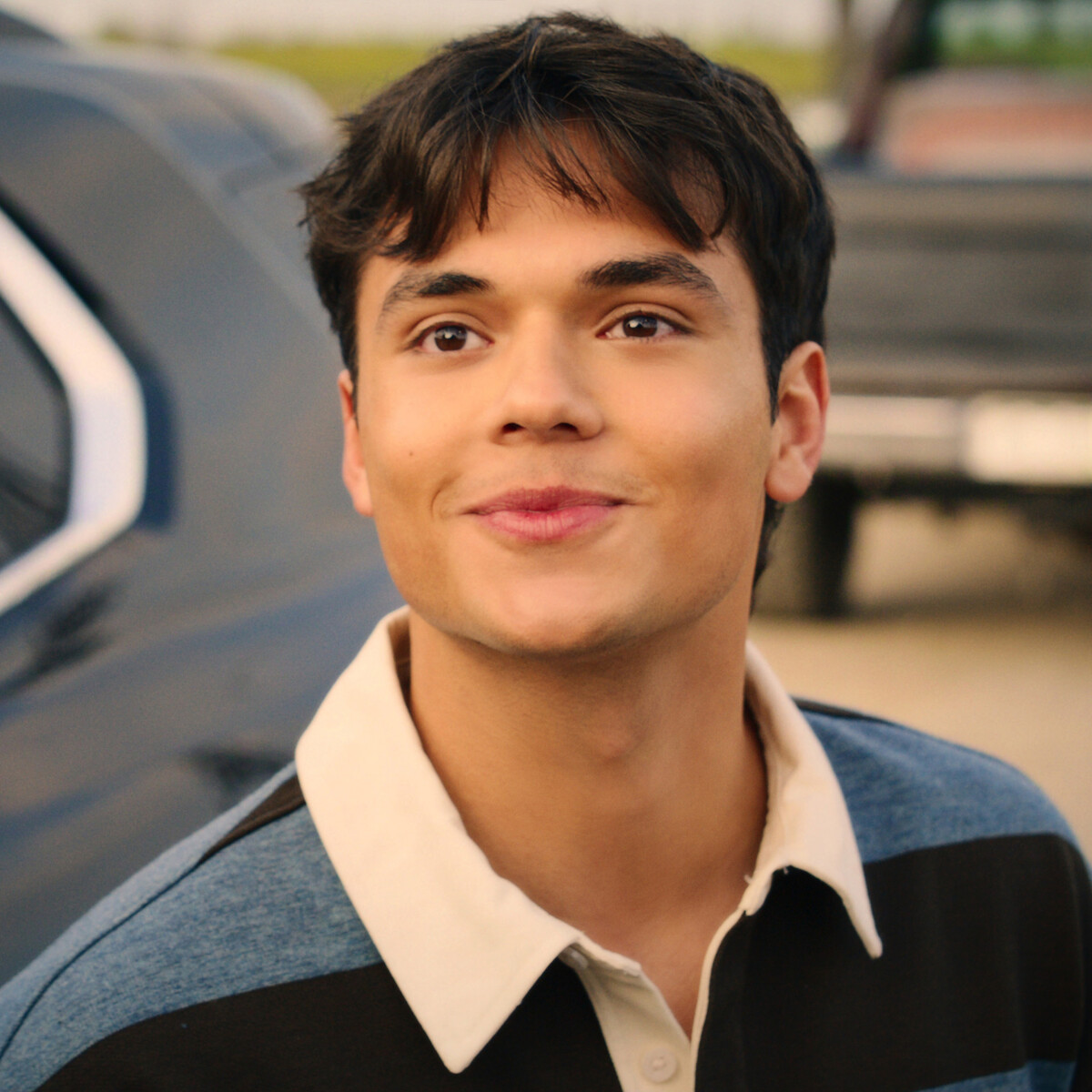 Young man smiling outdoors near a parked vehicle, wearing a blue and black striped collared shirt, with a blurred background suggesting a casual, possibly suburban or parking lot setting during daytime.