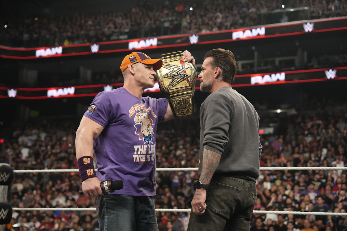 John Cena and CM Punk in a WWE ring face off, one holding a championship belt up to the other's face as a large audience watches in an arena setting.