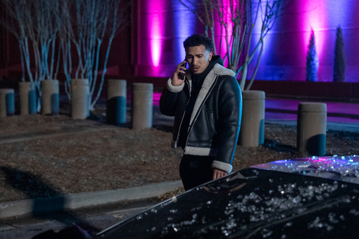 Xavier Smalls as Angel in a shearling jacket stands by a car on the phone at night in a parking lot, with colorful pink and blue lights illuminating leafless trees and a building in the background.
