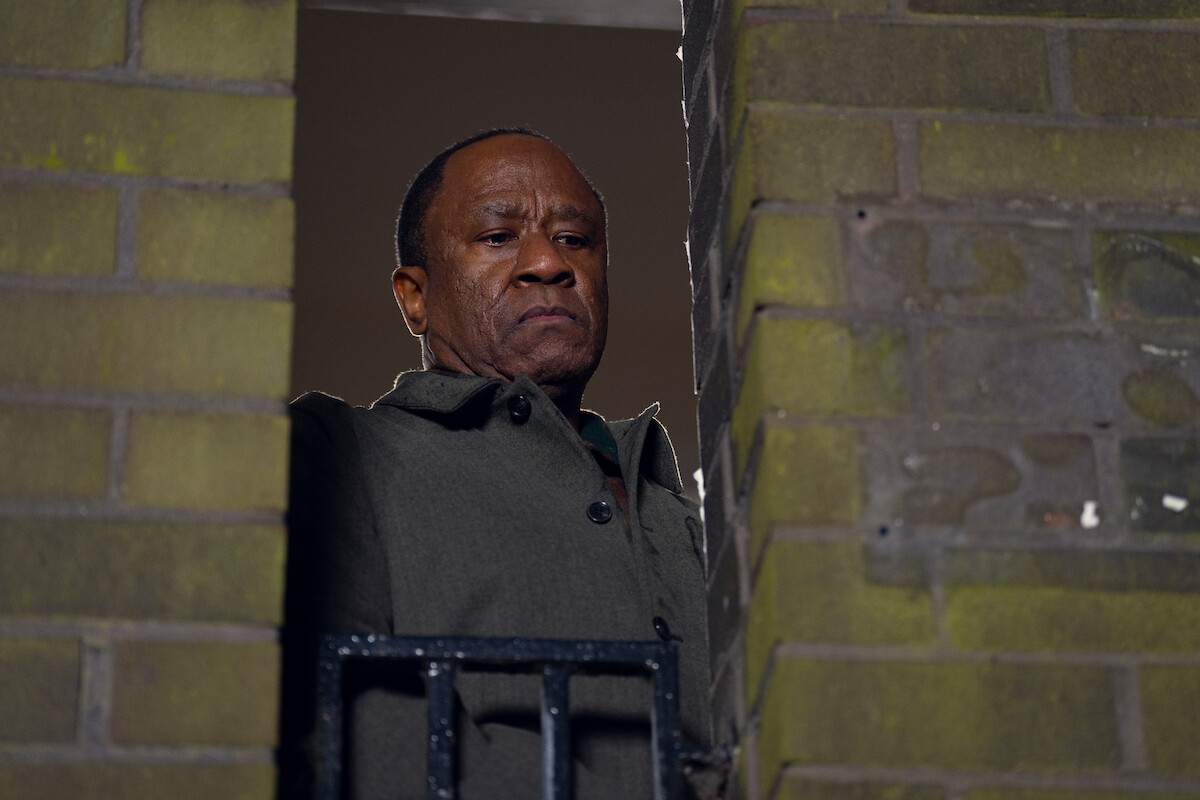 A serious man in a coat stands on a balcony between brick walls, looking down with a stern expression, suggesting a tense or contemplative mood in an urban setting.