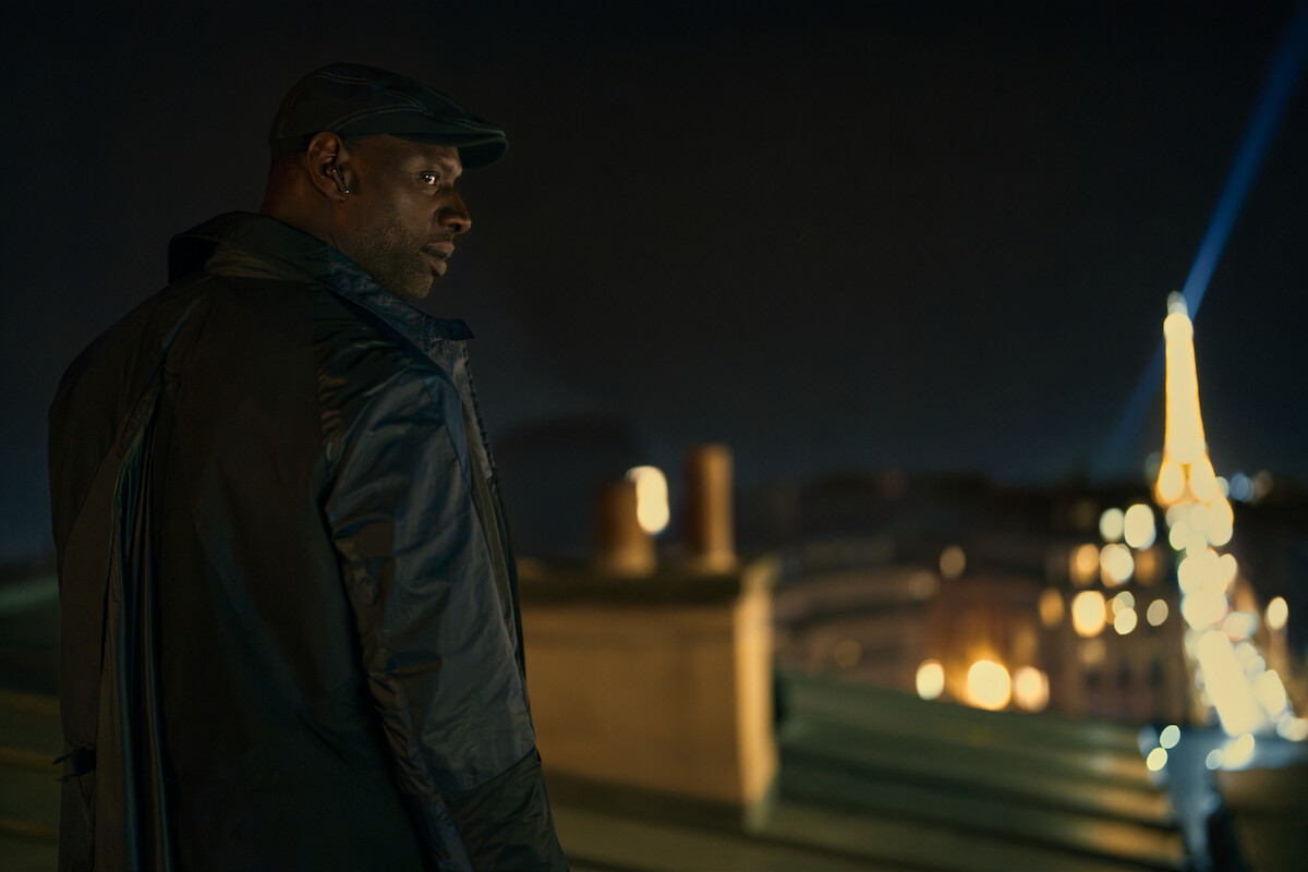 Um homem negro vestindo sobretudo observa a cidade de Paris do alto, com a torre Eiffel ao fundo
