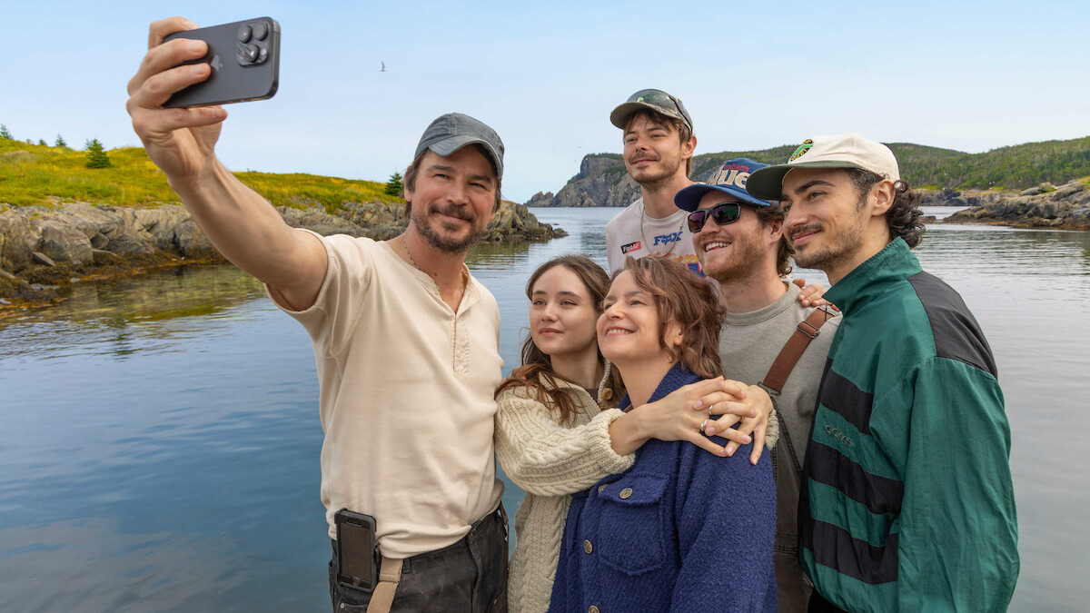 Six people take a group selfie by a scenic lakeside with rocky shores and greenery, dressed casually outdoors under a blue sky, all smiling and appearing happy together.