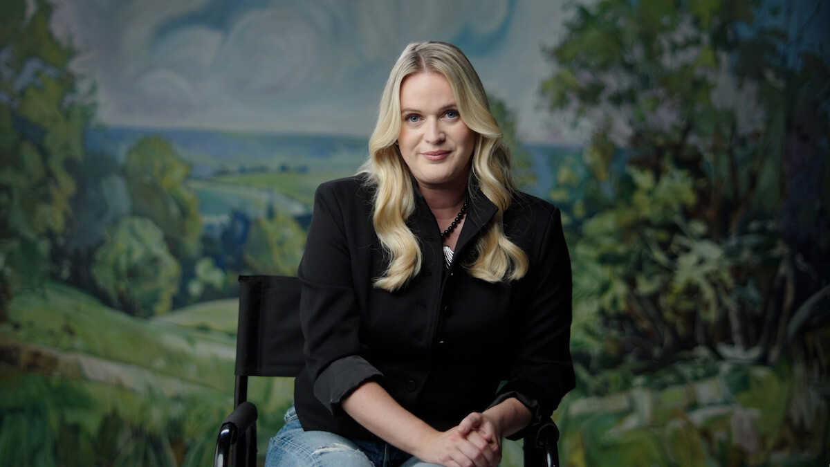 A woman with long blonde hair sits on a black chair, wearing a black blazer and jeans, in front of a painted backdrop depicting a green countryside landscape with trees and a cloudy sky.