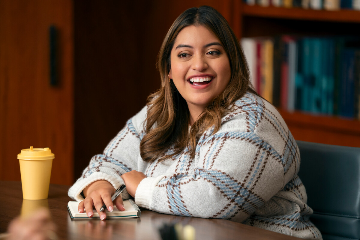Smiling woman in a cozy sweater sits at a conference table with a notebook and pen, a yellow coffee cup nearby, and bookshelves in the background, suggesting a meeting or discussion in an office or library setting.