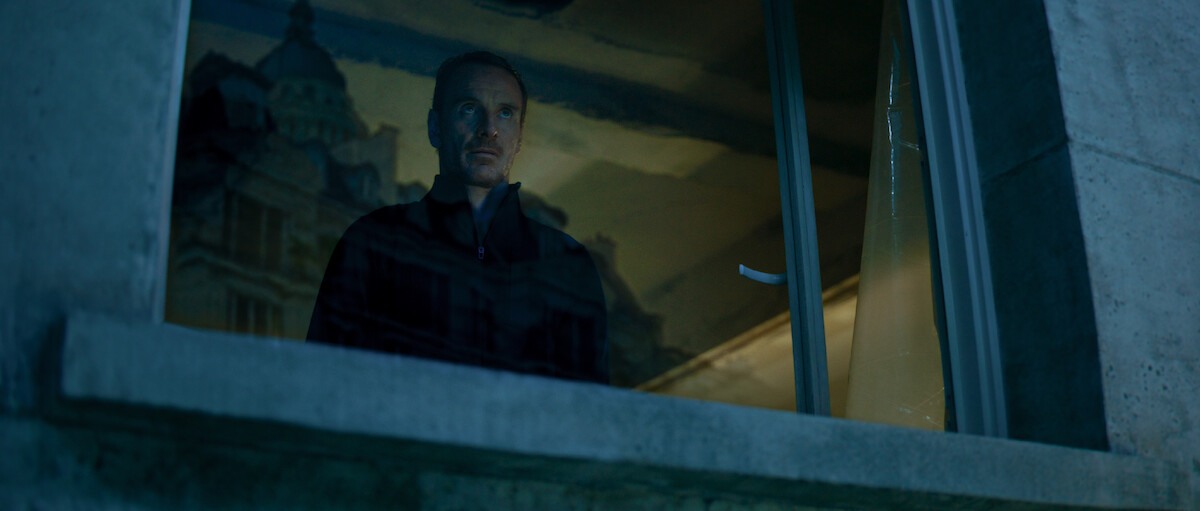 Michael Fassbender as The Killer wears all black and looks through a window in ‘The Killer’