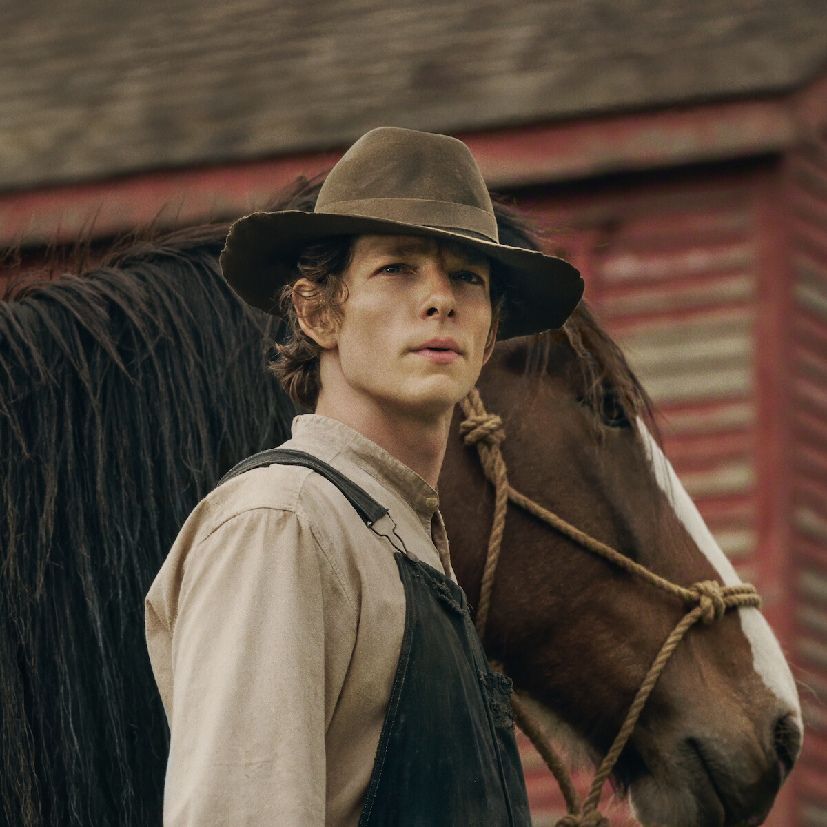 Mike Faist as Charles Trask in a brown hat and overalls stands next to a horse in front of a rustic red wooden building, evoking a rural farm or western setting.Mike Faist as Charles Trask in a brown hat and overalls stands next to a horse in front of a rustic red wooden building, evoking a rural farm or western setting.