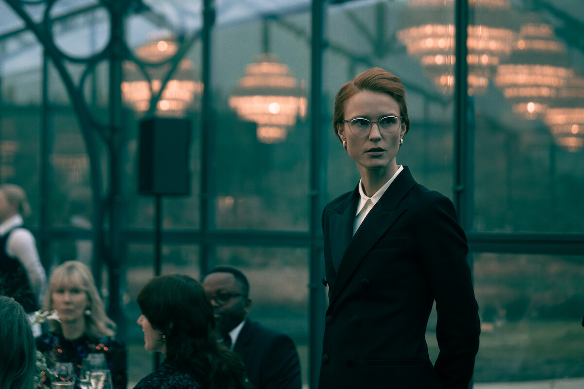 Amanda Collin wears an updo, glasses, and suit and while looking off into the distance with a serious expression.