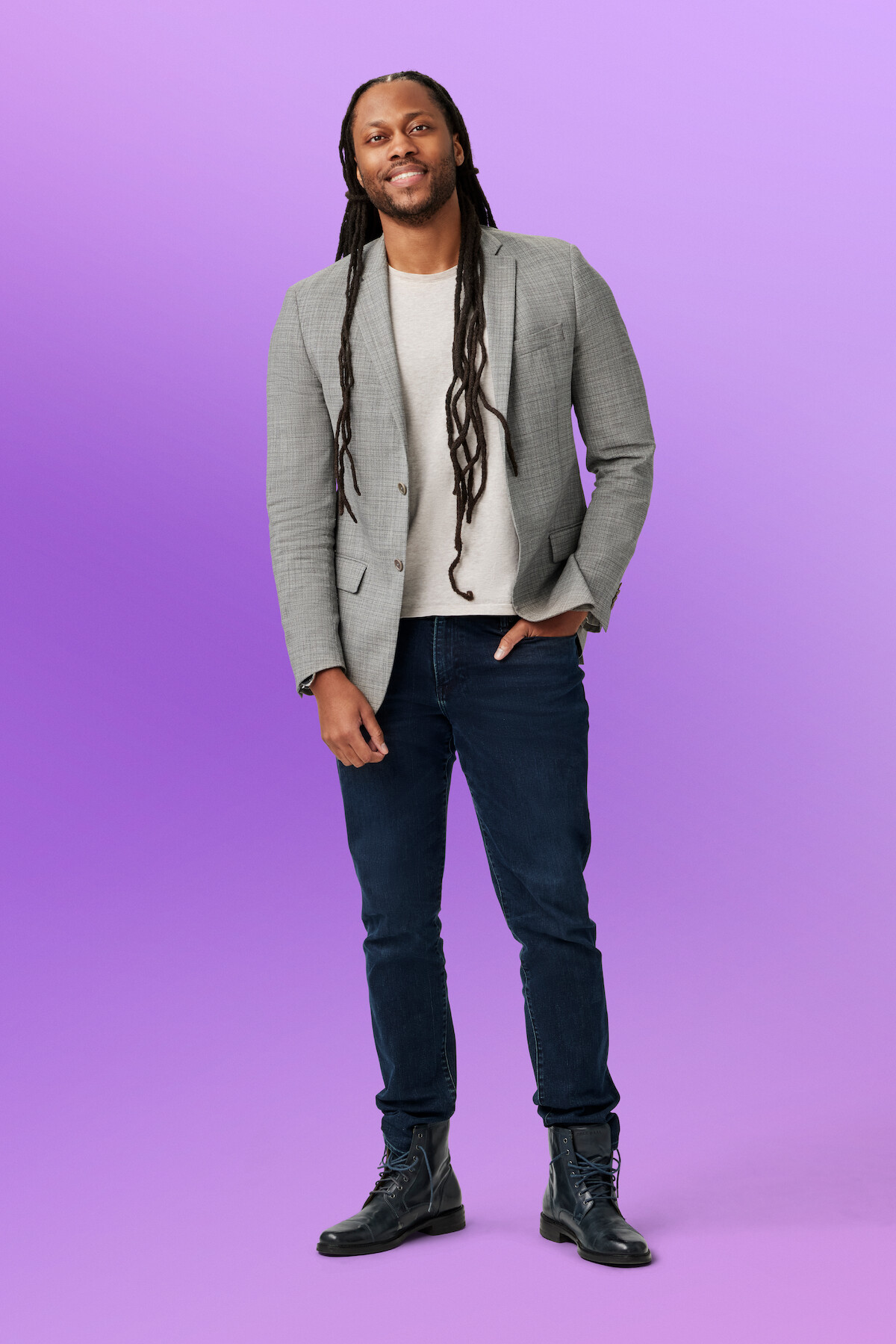 Vic wearing a gray blazer, light shirt, dark jeans, and boots stands smiling in a studio with a solid purple background.