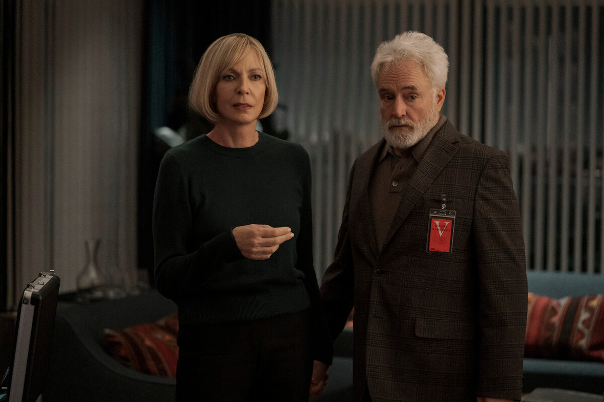 Two adults stand indoors in a modern room with striped curtains and a couch with red pillows; the woman has short blond hair and wears a dark sweater, while the man has gray hair, a beard, and a suit with a red ID badge.