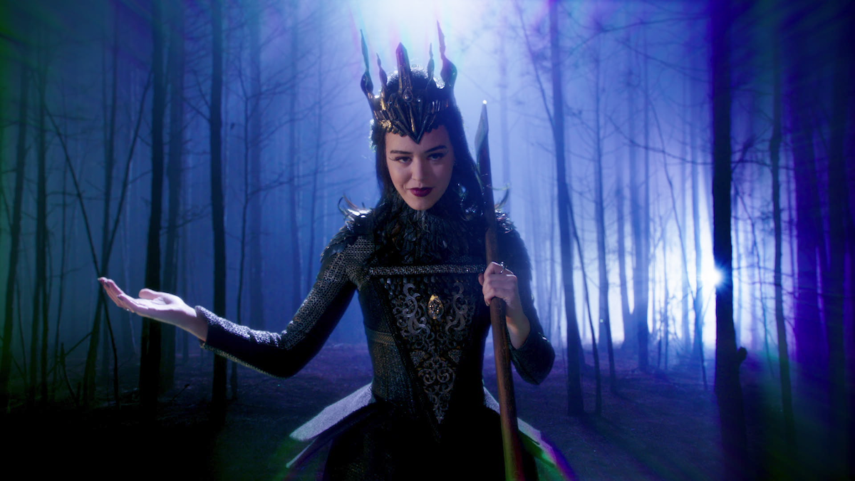 A woman in a dark, ornate crown and gown stands in a misty, eerie forest at night, holding a staff and gesturing dramatically, illuminated by blue and purple lighting.