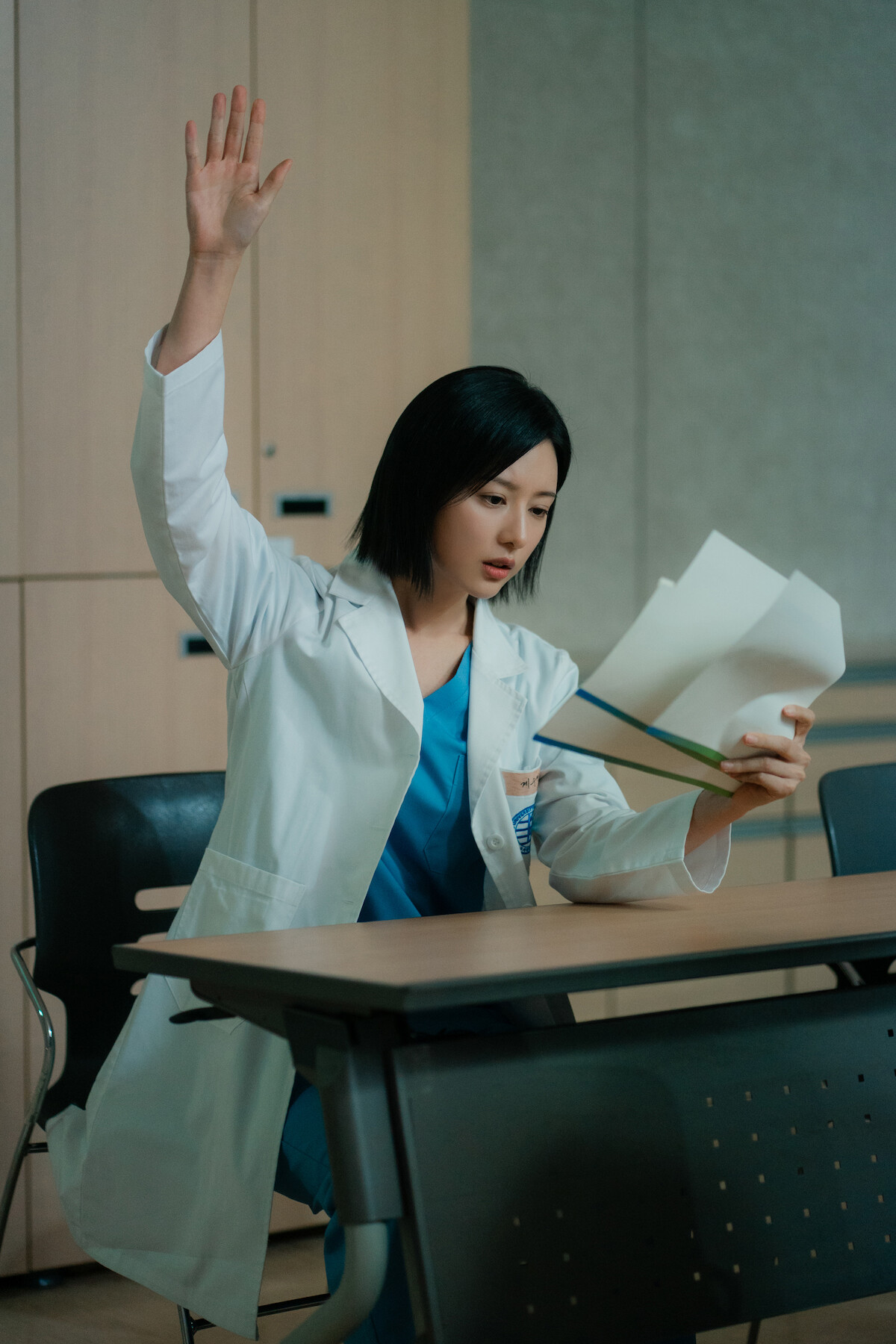 Uma mulher asiática de cabelos pretos e curtos levanta a mão enquanto lê um relatório médico. Ela está sentada numa mesa e veste jaleco branco e uma roupa interna azul, típica de hospitais