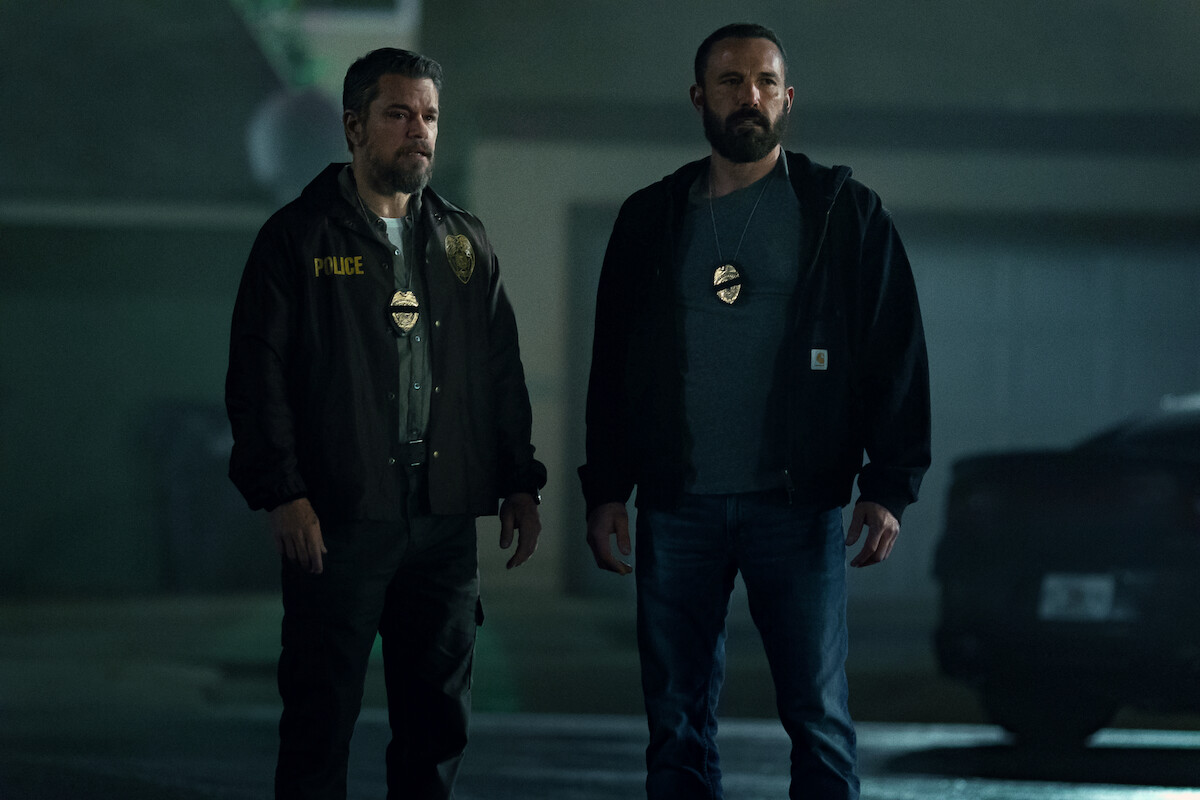 Two male police officers with badges stand in a dimly lit outdoor urban setting at night, near a car and a building, appearing serious and alert.