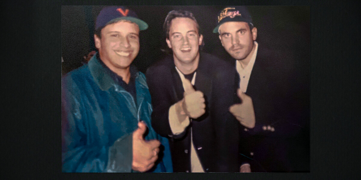 Three men posing together indoors at night, all smiling and giving thumbs up to the camera; two wearing baseball caps and jackets, standing in a casual social setting.