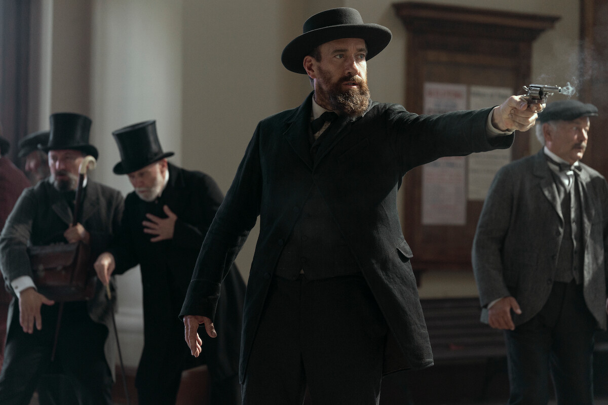 A bearded man in old-fashioned attire and hat points a revolver in a tense courtroom scene as alarmed men in Victorian clothing react in the background, creating a dramatic, suspenseful atmosphere.