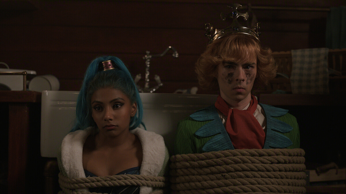Two people in colorful clothing, one with blue hair and the other with a crown, are tied up with rope in a dimly lit wooden room with a sink and various objects in the background.