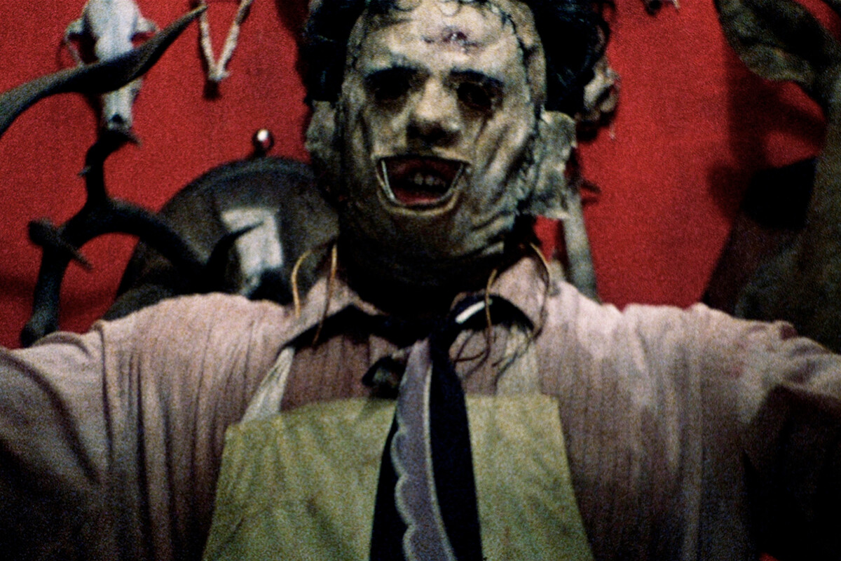 A person wearing a mask and apron stands with arms raised in a dimly lit, unsettling room decorated with animal skulls and red walls.