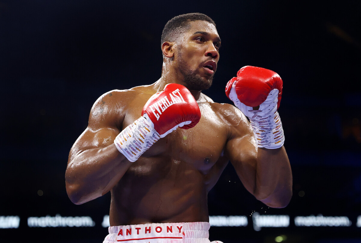 A focused male boxer in a brightly lit boxing ring, wearing red gloves and white shorts, appears ready to fight. The environment is energetic and intense, with a dark, blurred crowd and arena in the background.