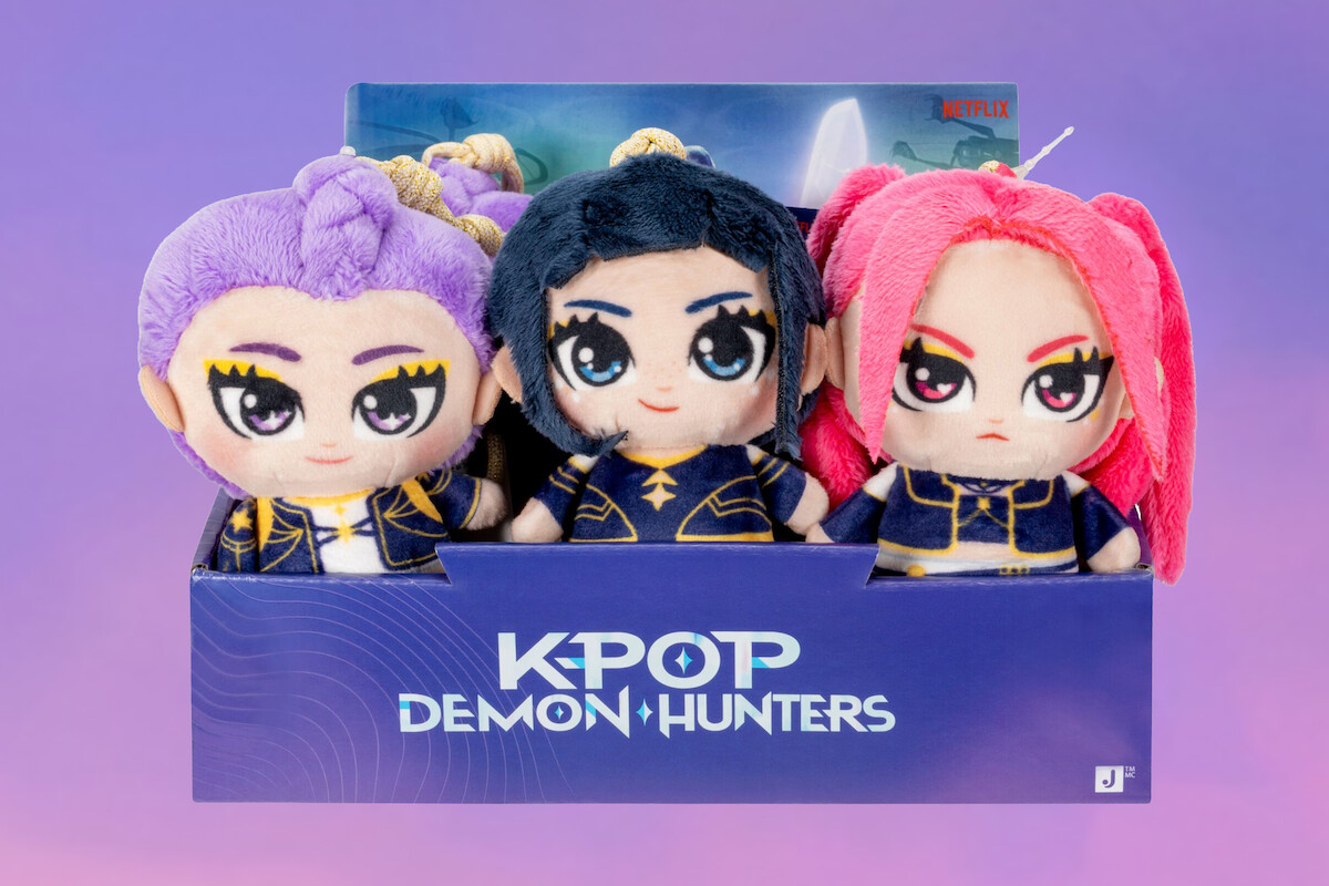 Three colorful K-pop Demon Hunters plush dolls with unique hair colors and outfits are displayed in a branded box against a pastel purple background.