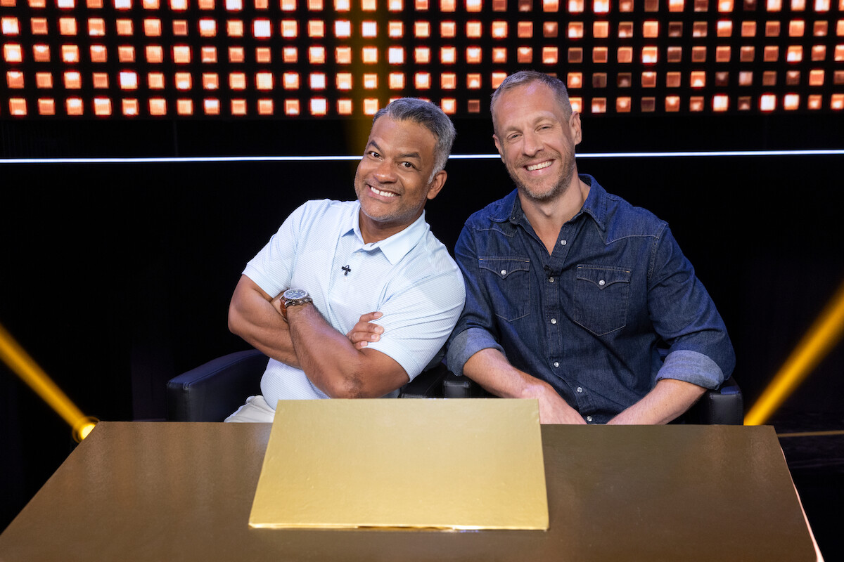 Two smiling men sit together at a table with a gold centerpiece in a brightly lit studio, creating a friendly and relaxed atmosphere.