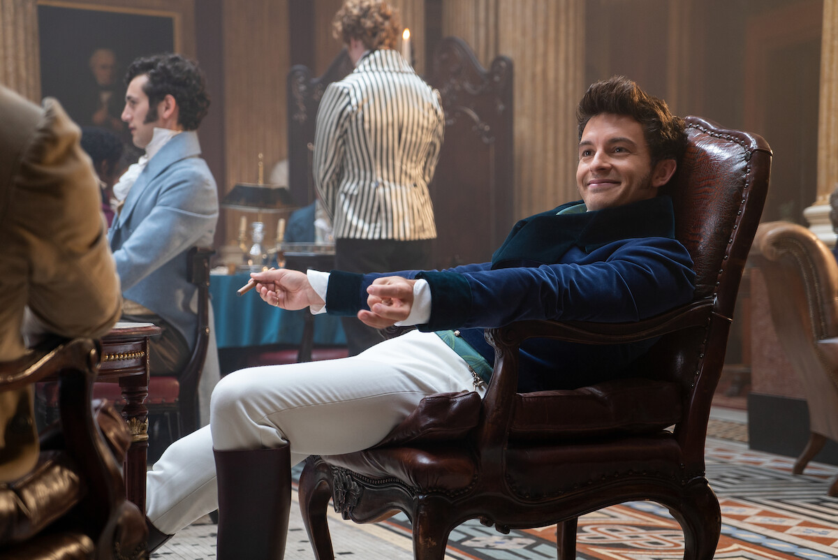 A smiling man in Regency-era clothing lounges in an ornate chair in a lavish room with columns, patterned floor, and other people in period attire, creating a historical social scene.