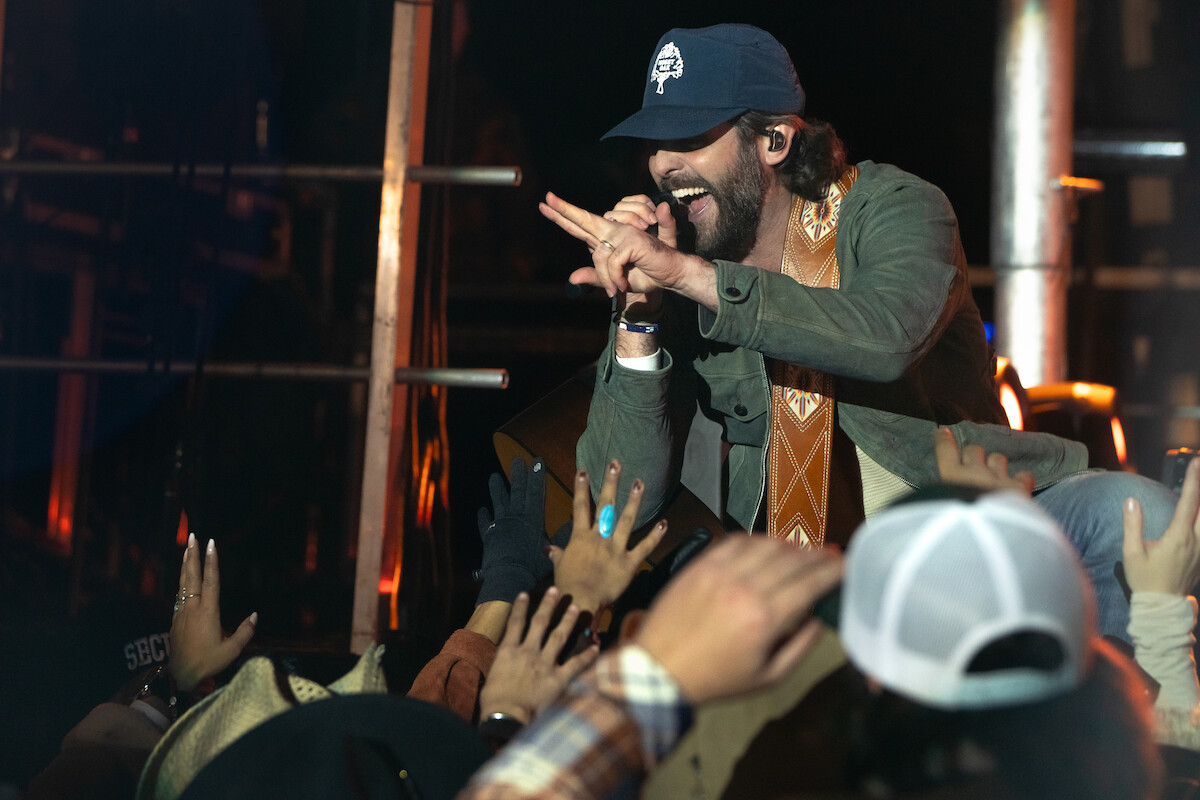 Male singer wearing a cap and casual jacket performs energetically on stage, reaching out to enthusiastic fans in the front row, with concert lighting and metal scaffolding visible in the background.