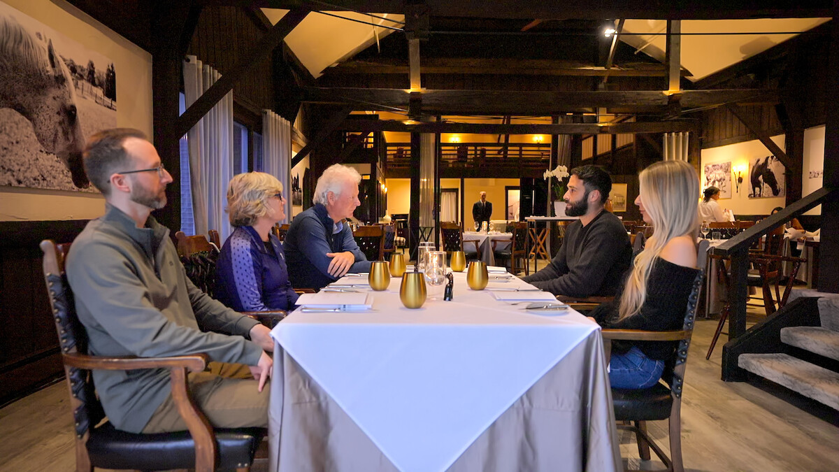 Cinco pessoas conversam sentadas à mesa em um restaurante rústico, de iluminação acolhedora, com vigas de madeira, toalha de mesa branca, taças douradas e fotos de grandes animais nas paredes.