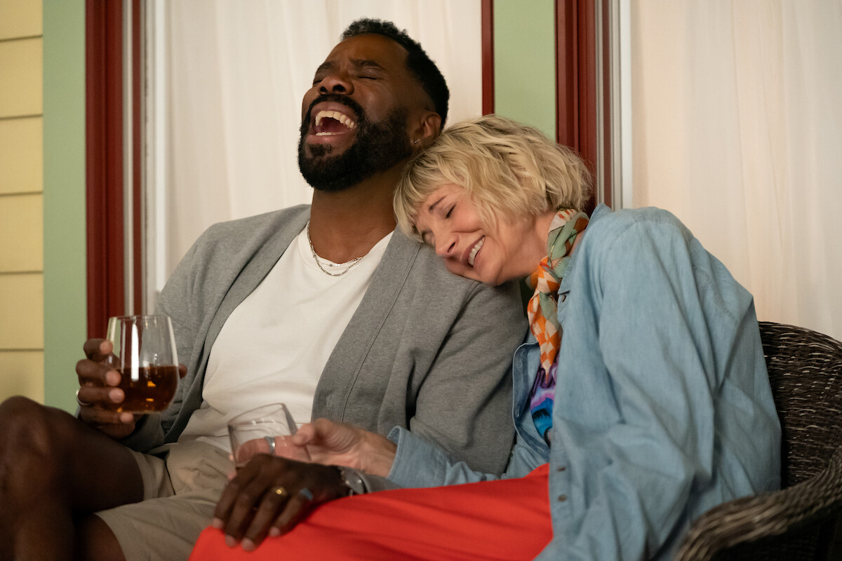 Two people sitting closely on a porch, sharing a joyful moment with drinks in hand. One person laughs heartily while the other rests their head on his shoulder, looking content. Cozy, relaxed evening setting.