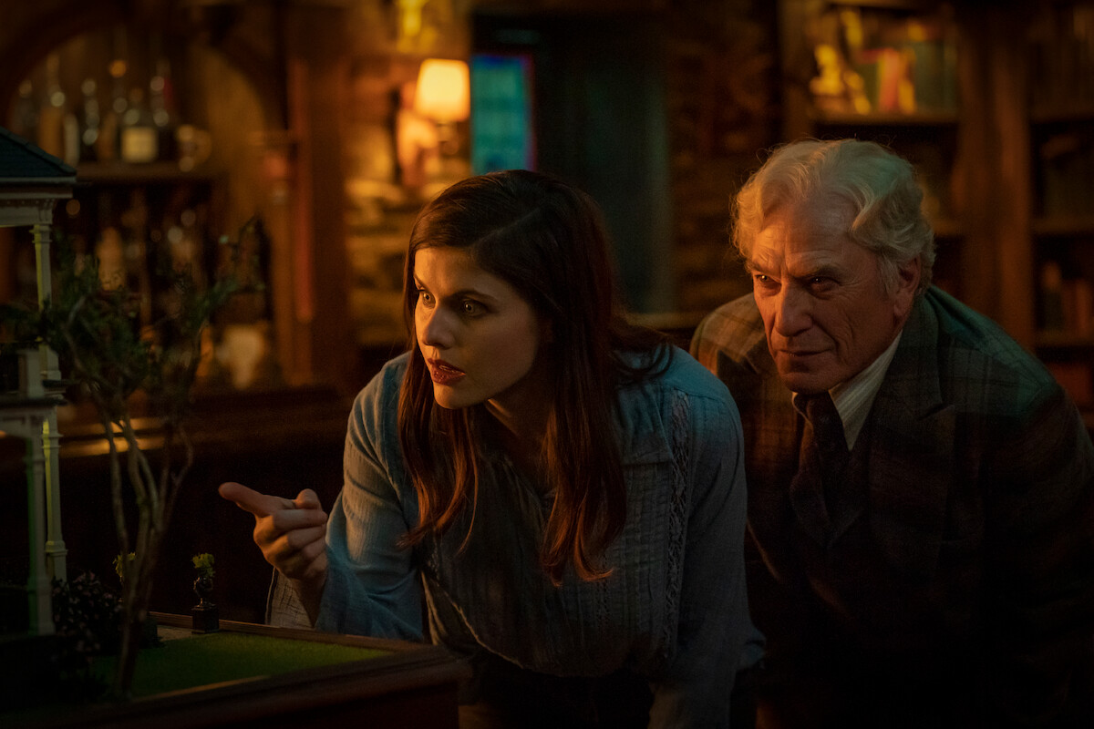 A woman and an older man examine a detailed miniature house indoors, surrounded by warm lighting, bookshelves, and a cozy, wood-accented room, suggesting curiosity or investigation.