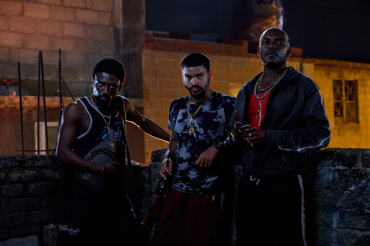 Three men with serious expressions stand behind a rough brick wall at night in an urban outdoor setting, wearing casual streetwear and jewelry, dimly lit by artificial lighting.