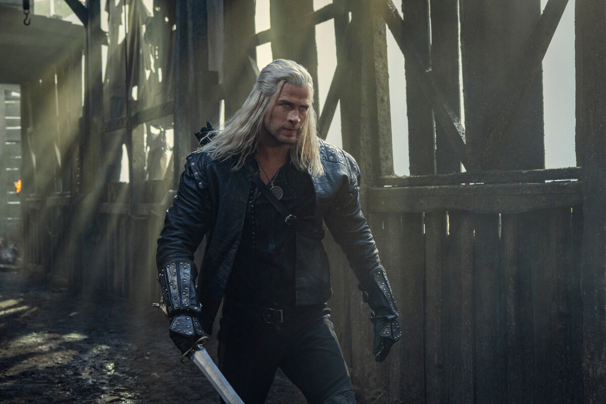 A man with long white hair in black armor walks through a dimly lit, rustic wooden structure, holding a sword, with sunlight filtering through, creating a tense and dramatic medieval atmosphere.