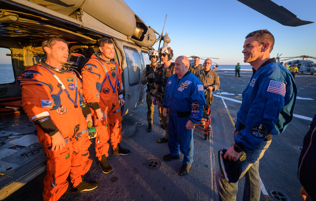 Astronauts in orange and blue suits stand on an aircraft carrier flight deck by a helicopter at sunset, with crew members and equipment visible, preparing for or after a mission.