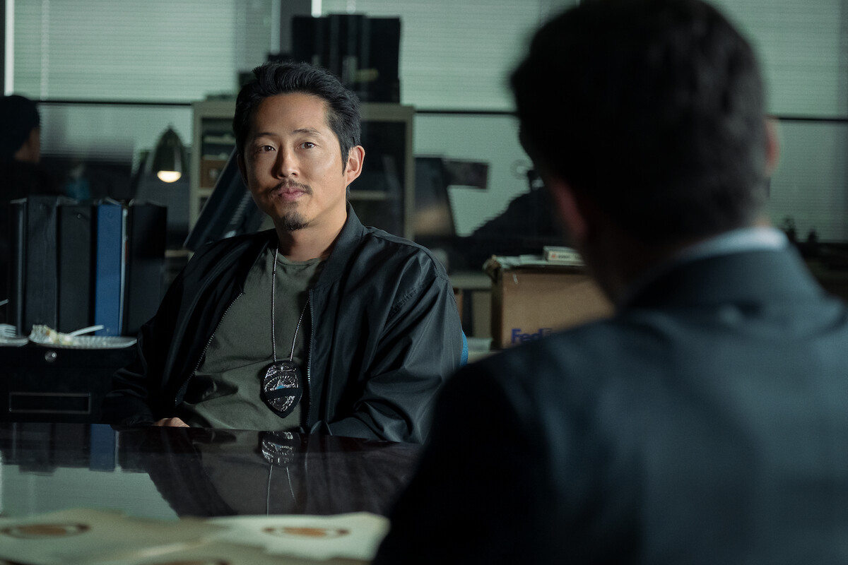A man with a badge sits at a desk in an office setting, facing another man whose back is to the camera. There are files, office supplies, and boxes in the background, indicating a workplace or police station environment.