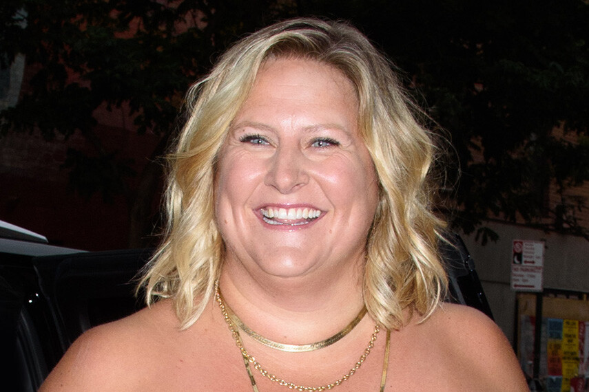 A woman with blonde hair smiling outdoors in an urban setting at night, wearing layered necklaces, with a joyful and cheerful mood. Trees and street signs are visible in the background.