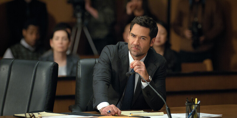 A serious man in a suit sits at a desk in a courtroom, speaking into a microphone. Papers are spread in front of him, with people and cameras in the background, creating a tense, formal atmosphere.