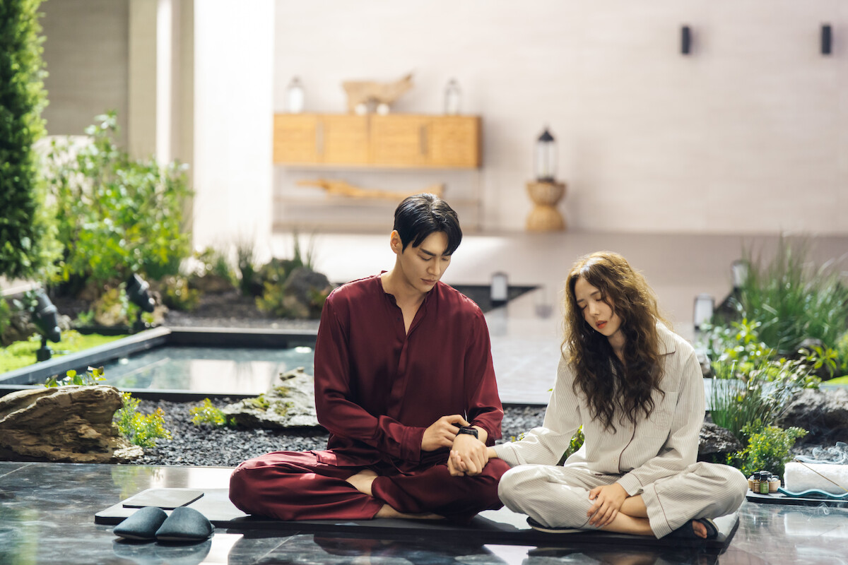 Um casal sul-coreano está sentado sobre um jardim interno. O homem veste um pijama de seda vinho, e a mulher, um pijama bege. Eles estão de mãos dadas