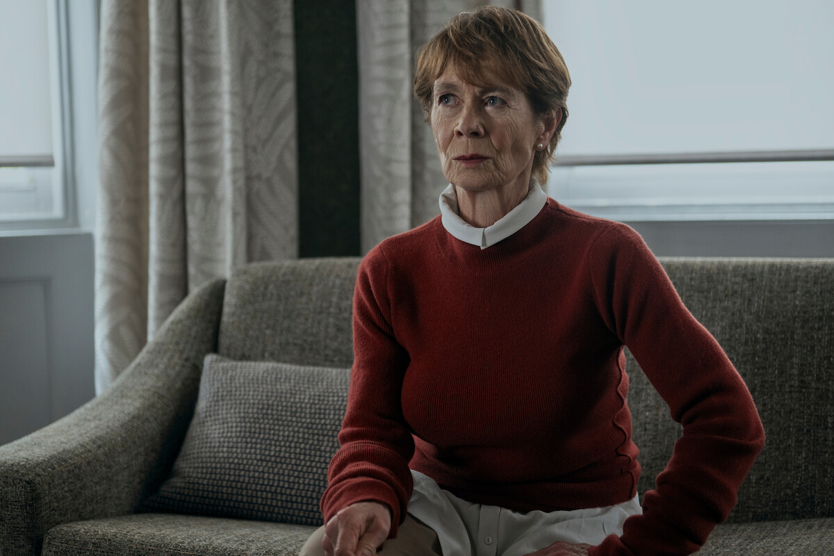 An older woman with short hair sits on a gray sofa in a softly lit room with large windows and patterned curtains, wearing a red sweater over a white collared shirt, looking contemplative.