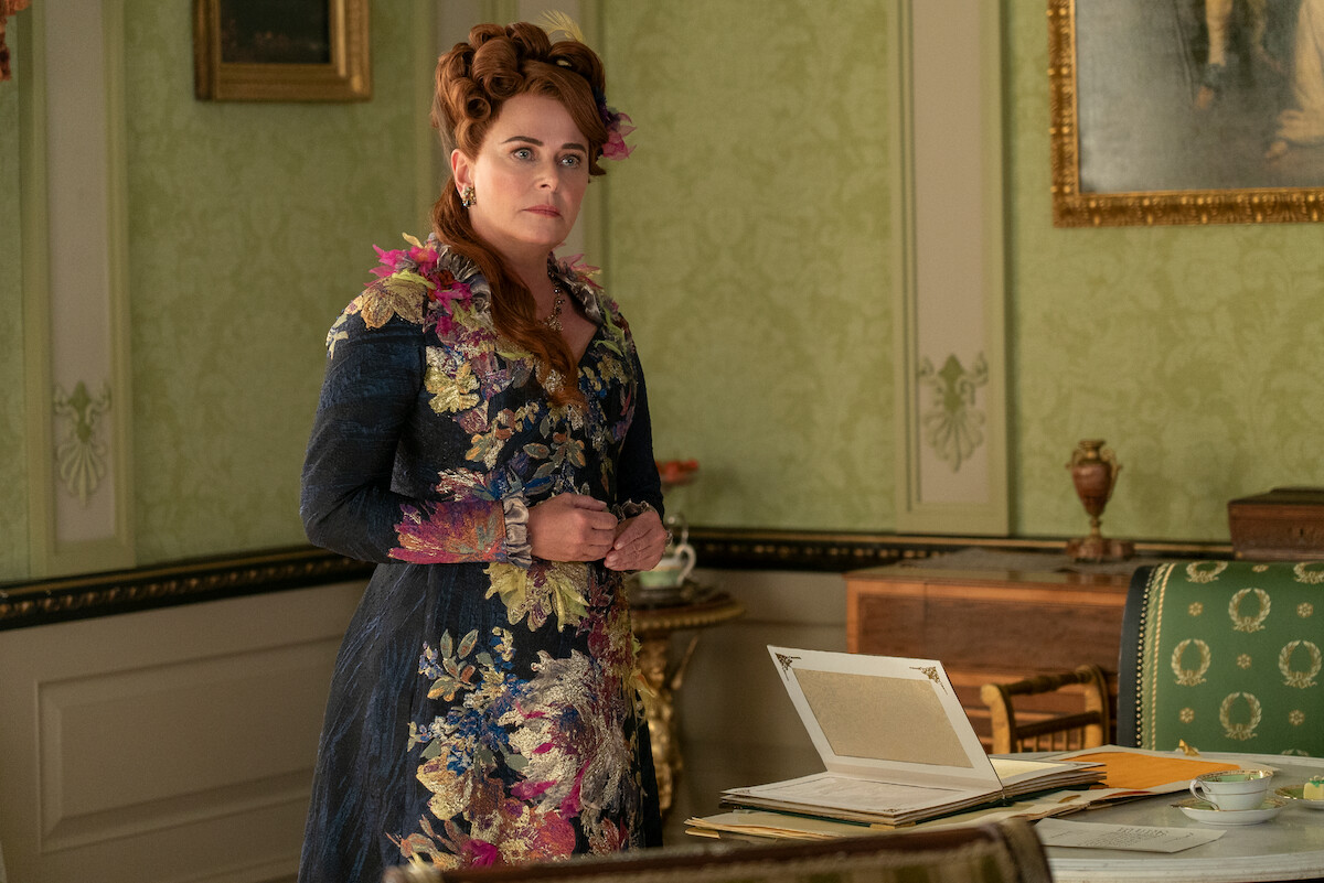 A woman in an ornate floral gown stands in an elegant, vintage-style room with green wallpaper, framed art, and a desk with papers and an open box, suggesting a historical or period setting.