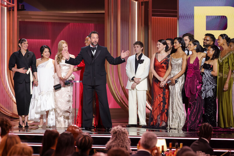 A group of people in formal attire stand on stage at an awards ceremony, with one man speaking at a microphone and others gathered around him, surrounded by a decorated, well-lit event venue with an audience visible in the foreground.