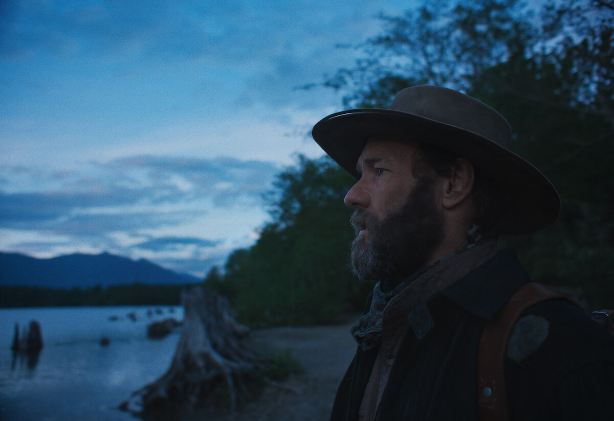 Joel Edgerton looks into the distance with a sunset and tress behind him.