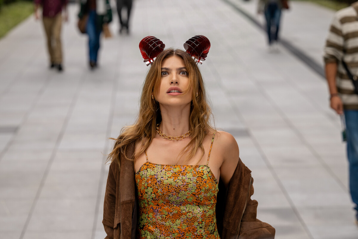 Uma mulher branca de cabelos castanhos veste um vestido estampado de flores alaranjadas e um acessório no cabelo. Ela olha para o alto com uma expressão preocupada
